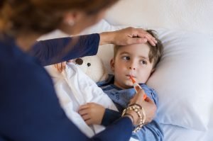 what to do when your child has a fever; Sick boy with thermometer laying in bed and mother hand taking temperature. Mother checking temperature of her sick son who has thermometer in his mouth. Sick child with fever and illness while resting in bed.