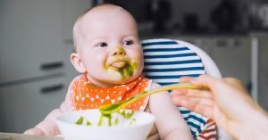 Feeding. Messy smiling baby eating with a spoon in high chair. Baby's first solid food. Mother feeding little child with spoon of puree. Daily routine. Finger food. Healthy child nutrition; blog: Baby's First Foods: Tips for Starting Solids