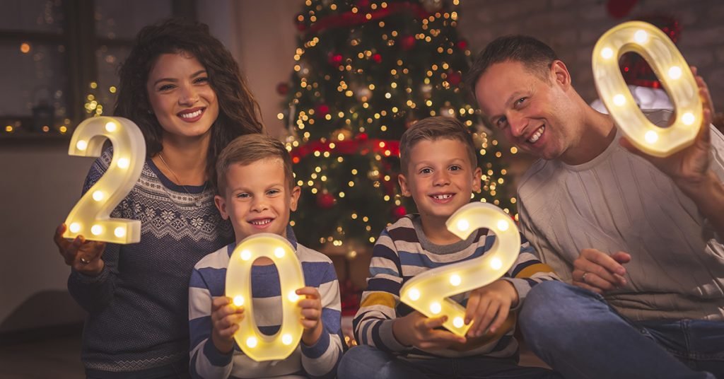 Parents celebrating New Years Eve at home with kids, sitting by the Christmas tree, holding illuminative numbers 2020 representing the upcoming New Year; blog: 9 New Year’s Resolutions for the Whole Family
