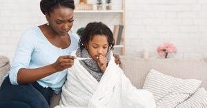 Flu Concept. Worried black mother checking her daughter's temperature, child covered in blanket; blog: Is My Child Too Sick for School?