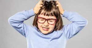 irritated 6-year old young girl with eyeglasses pulling out her hair for itchy allergies or lice or scratching her head for nervous disagreement, grey background; blog: A Parent’s Guide to Head Lice Prevention