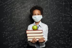Happy child in medical protective face mask holding books and green apple on blackboard background; blog: Face Mask Myths Debunked