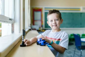 Young boy playing with a blue toy truck at a classroom table by a window.