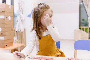 A young girl in a mustard dress plays with a puzzle at a preschool wooden table indoors.